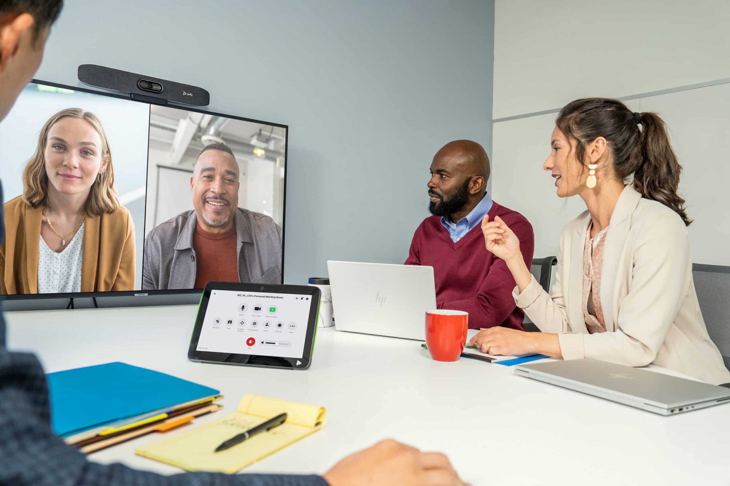 In an office meeting, 2 on screen, 2 at a table with laptops/tablet. One points at the screen.