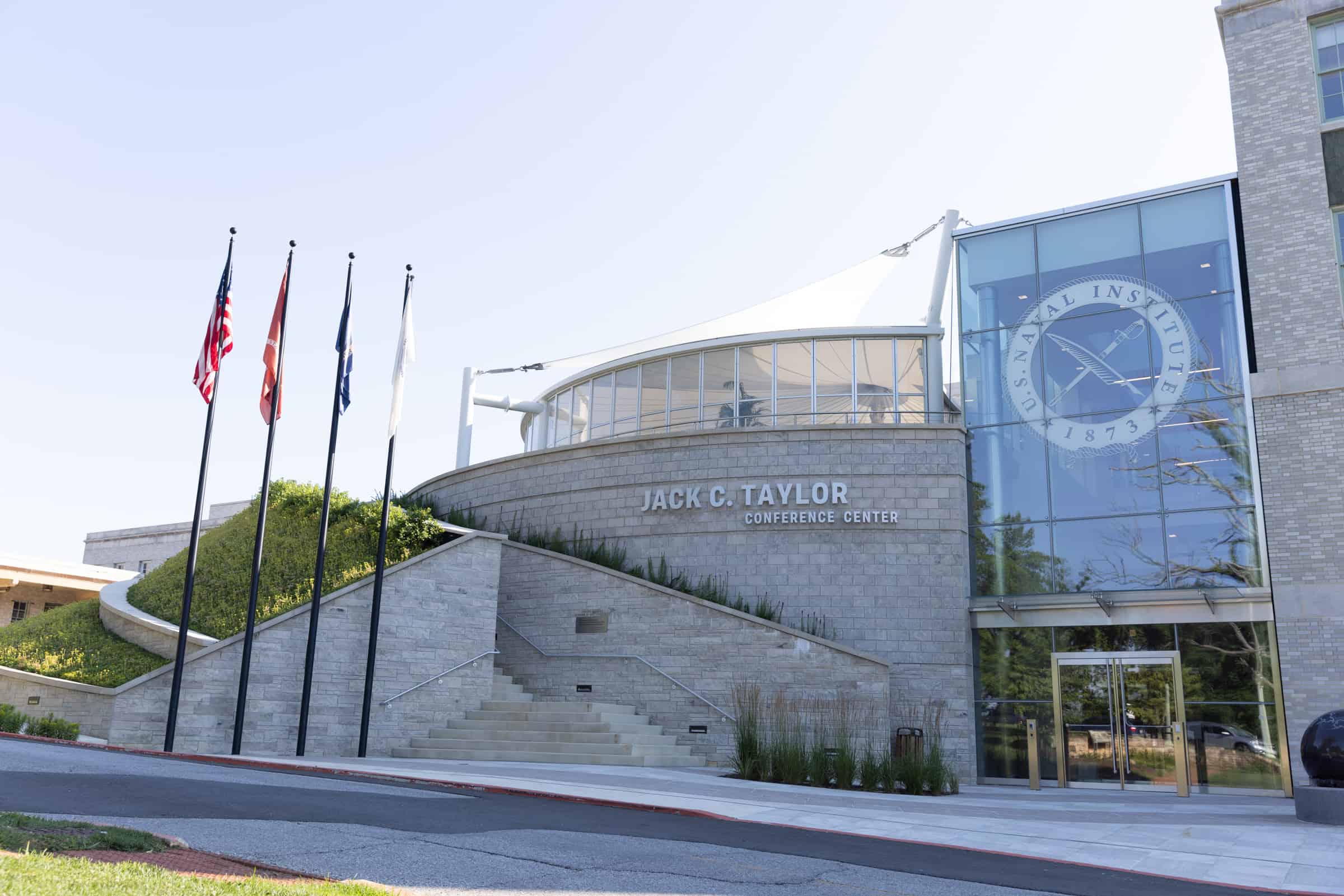 Modern circular design with large windows, four flags by entrance stairs, and Naval Institute 1873 logo on Jack C. Taylor Center.